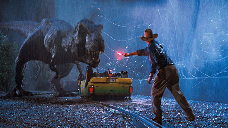 Sam Neill as Alan Grant in Jurassic Park, standing in the rain and waving a flare while facing down a Tyrannosaurus Rex, approaching next to a turned over car.