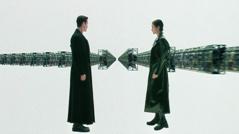 Keanu Reeves as Neo and Carrie-Anne Moss as Trinity in The Matrix, facing each other in a white room filled with rows of computer machinery.