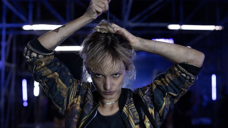 Agathe Rousselle as Alexia/Adrien in Titane, looking toward the camera and putting a pin in her hair in an industrial-looking club.